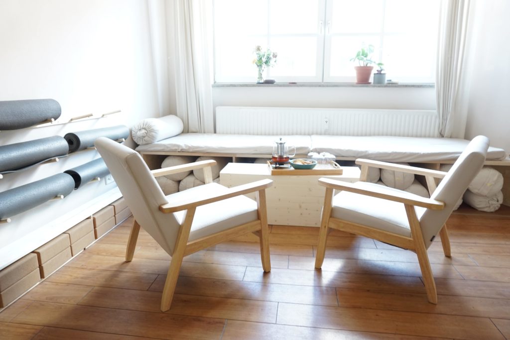 Eine gemütliche Sitzecke im großen Yogaraum mit einer Holzbank, Sitzkissen und sanfter Dekoration. Helles Tageslicht fällt durch die großen Fenster.